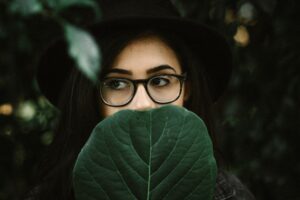 artistic photo of young woman with dark hair and glasses holding a large dark green leaf over the lower half of her face