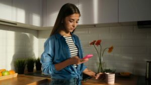 a young woman standing in a kitchen looking at her mobile phone